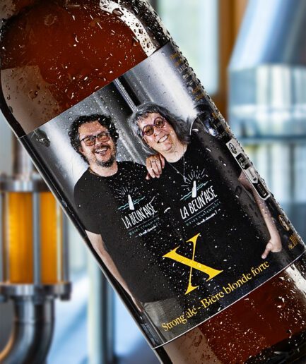 Montage d'une bouteille en diagonale de la bière X de la brasserie La Beun'aise, avec en arrière-plan du matériel de brasserie (tuyauterie inox, cuves, mireur). L'étiquette de la bière présente les deux fondateurs de la brasserie, côte à côte, portant un t-shirt noir avec le logo de la brasserie La Beun'aise ; ils se tiennent debout devant leurs cuves de fermentation. En surimpression de couleur orange, le nom de la bière, X, suivi du descriptif : Strong ale - bière blonde forte.